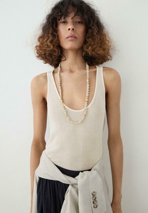 Donna con capelli ricci che indossa una canotta beige, una lunga collana di perline e una giacca leggera annodata in vita.
