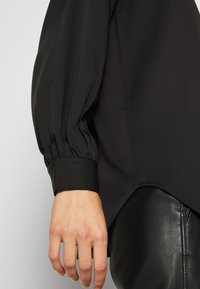 Black blouse featuring puffed sleeves, a smooth texture, and a fitted cuff. Hemline is curved and matches with slim black leather pants.