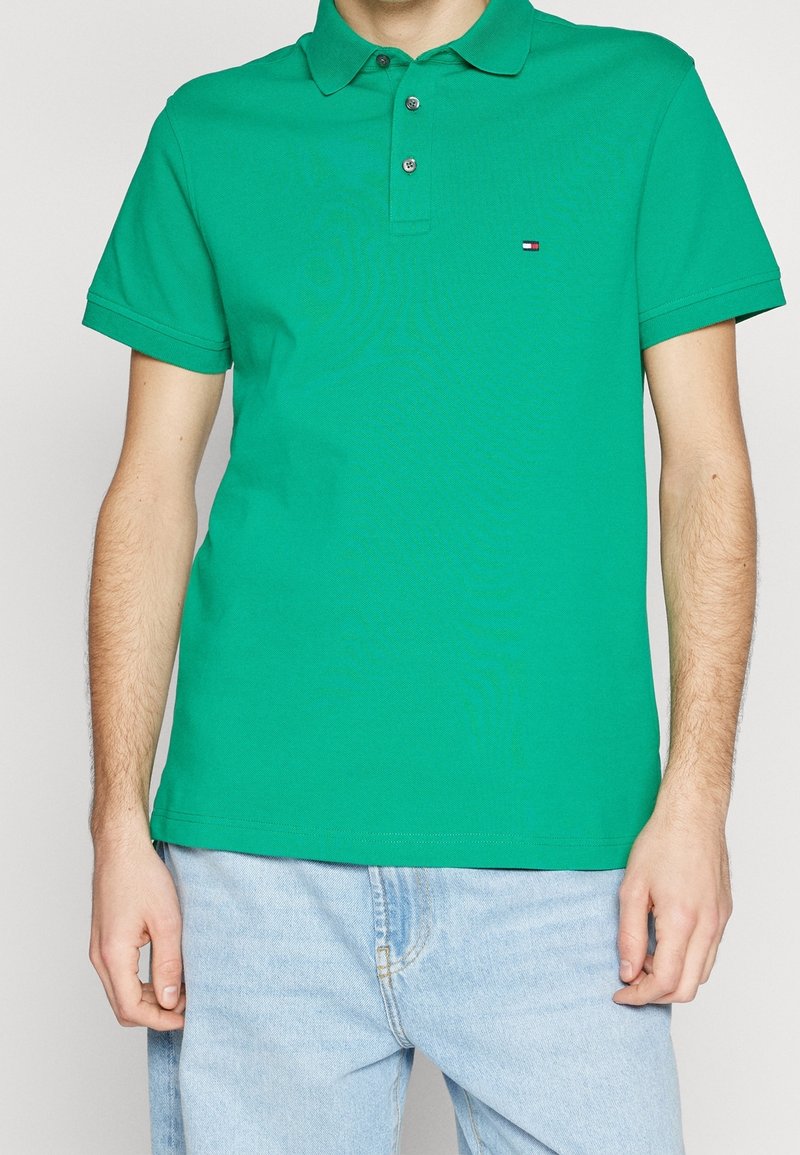 Homme portant un polo à manches courtes vert vif avec un petit logo sur la poitrine et un jean bleu clair, sur un fond uni.