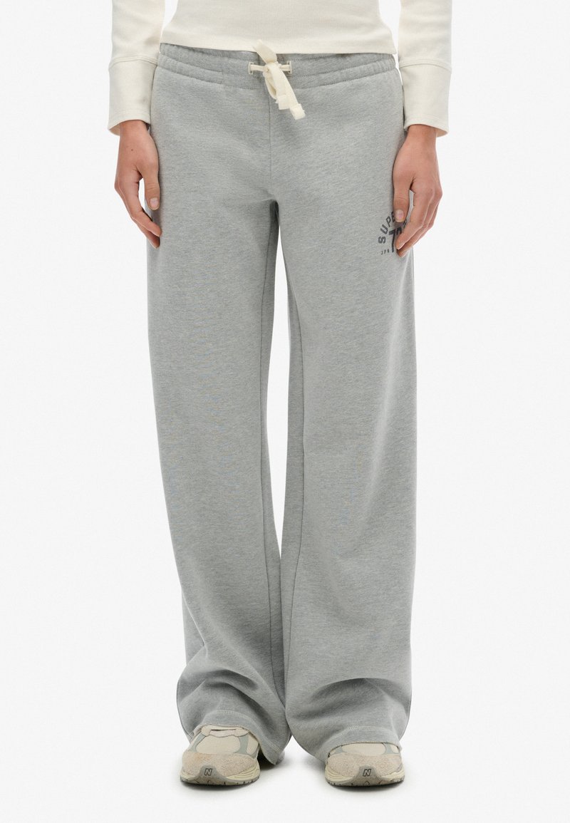 Persona que lleva pantalones de chándal gris holgados con cintura con cordón y zapatillas beige, de pie contra un fondo blanco.