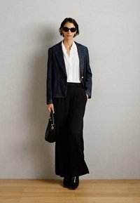 Kvinne med mørke solbriller, mørk blazer, hvit bluse, vide svarte bukser, svarte støvler, holder en svart håndveske mot en ensfarget vegg.