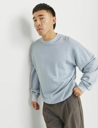 Svietlo modrý crewneck mikina vyrobená z mäkkej tkaniny s dlhými rukávmi, spárovaná so šedými nohavicami. Obe kusy majú voľný strih.