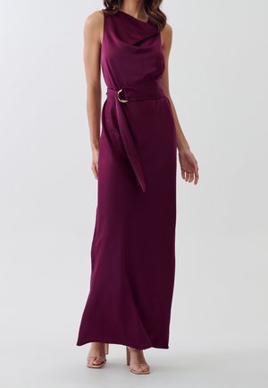 Mujer con vestido largo sin mangas de color púrpura oscuro con escote drapeado y cinturón a juego, combinado con sandalias beige de tacón.