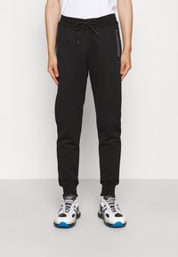 Schwarze Jogginghose mit einem Kordelzug am Bund, zwei Reißverschlusstaschen mit farblichen Akzenten und schmal zulaufenden Beinabschlüssen. Getragen mit sportlichen weißen und blauen Sneakers.
