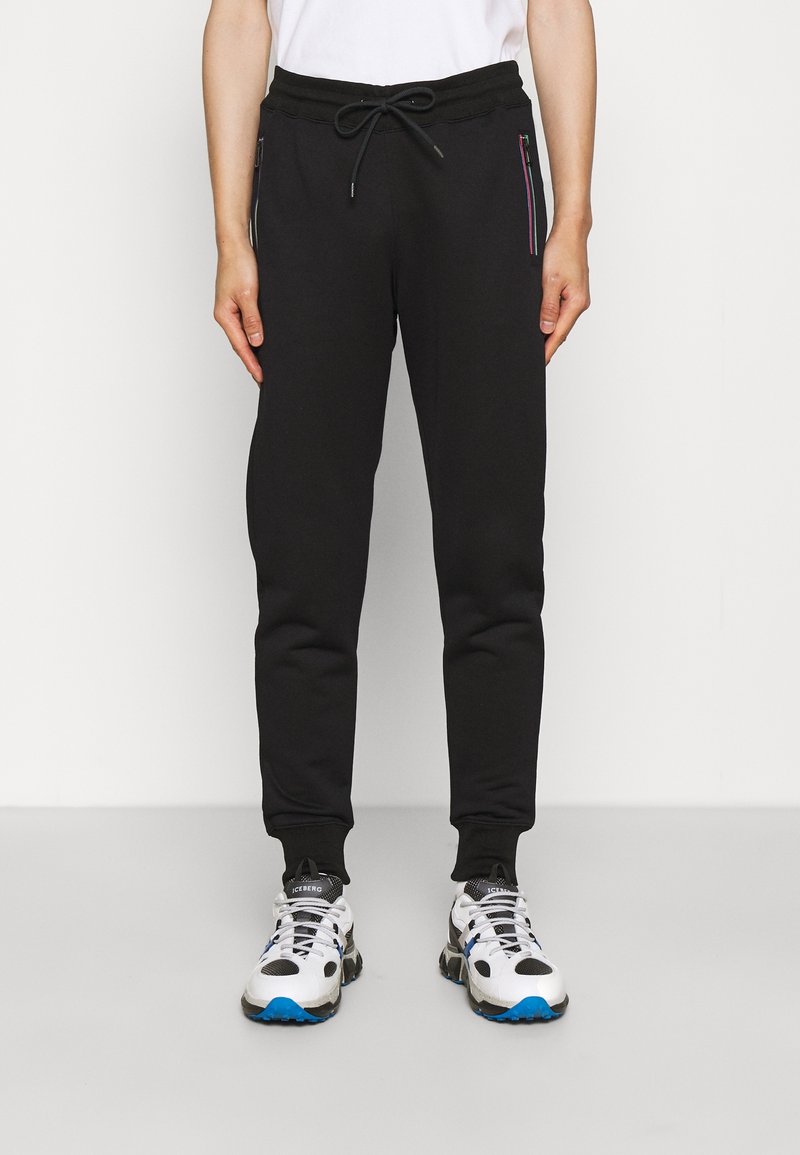 Schwarze Jogginghose mit einem Kordelzug am Bund, zwei Reißverschlusstaschen mit farblichen Akzenten und schmal zulaufenden Beinabschlüssen. Getragen mit sportlichen weißen und blauen Sneakers.