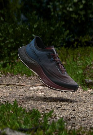 Buty do biegania trailowego Columbia w czarno-bordowym kolorze nad ziemną ścieżką z trawą, pokazujące bieżnik i logo marki na boku i języku.