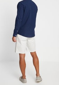 Marinblå långärmad skjorta, vita bomullsshorts, beige skor. Skjortan har manschetter med knappar och detaljer vid ryggen. Shortsen är knälånga.