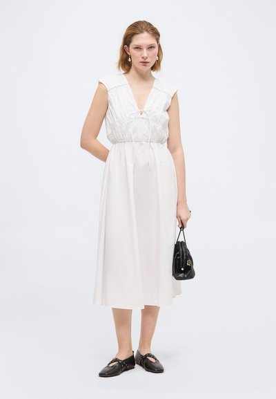 Femme portant une robe midi blanche sans manches tenant un petit sac à main noir, associée à des chaussures plates noires, debout devant un fond uni.