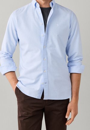 Mann trägt hellblaues Hemd mit Knöpfen über einem schwarzen T-Shirt mit hochgekrempelten Ärmeln und dunkelbrauner Hose vor schlichtem Hintergrund.