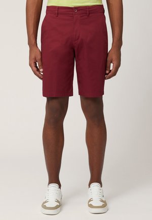 Burgunderrote Shorts mit flacher Vorderseite, Gürtelschlaufen und Seitentaschen; hergestellt aus leichtem Stoff. Kombiniert mit weißen Sneakern mit beigen Akzenten.