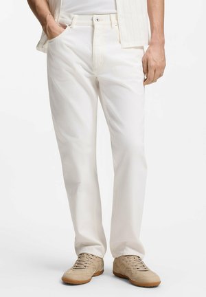 Mann trägt helle gerade geschnittene Hose, beige Sneakers und ein weißes Tanktop mit einem teilweise sichtbaren gestreiften Hemd.