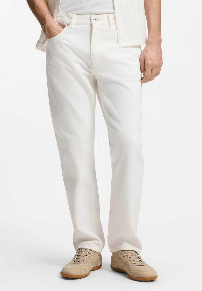 Homme portant un pantalon droit beige clair, des baskets beiges et un débardeur blanc avec une chemise rayée partiellement visible.