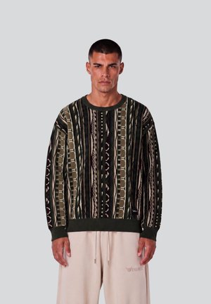 Mann trägt einen mehrfarbigen vertikal gestreiften Pullover und hellbeige Hose und steht vor einem einfarbig grauen Hintergrund.