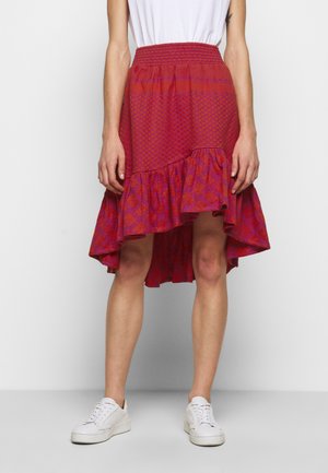 Mujer con una falda asimétrica con volantes en tonos rojo y púrpura, combinada con zapatillas blancas y una camiseta blanca metida por dentro, sobre un fondo liso.