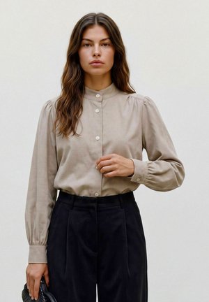 Femme aux longs cheveux bruns, portant un chemisier beige boutonné et un pantalon noir, tenant un sac à main noir sur un fond clair uni.