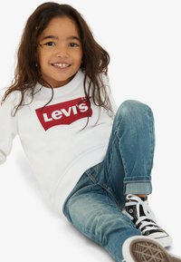 Λευκή μπλούζα με κόκκινο λογότυπο Levi's, συνδυασμένη με μπλε τζιν παντελόνι και μαύρα sneakers. Παιδί καθιστό, με μακριά και κυματιστά μαλλιά.