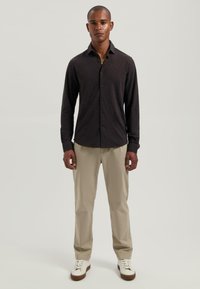 Man staat met het gezicht naar voren, gekleed in een donkerblauwe lange mouwen overhemd, beige broek en witte sneakers, tegen een effen lichte achtergrond.