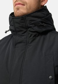Black jacket with a high collar, featuring drawcords and a smooth, matte texture. Includes a patch pocket with a button detail.