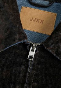 Zwarte corduroyjas met een zilveren rits, voorzien van een bruine leren label met de tekst "JJXX" en een blauwe denim voering.