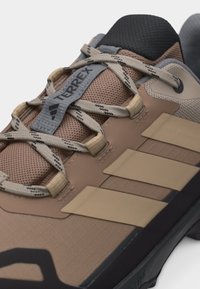 Κοντινό πλάνο ενός καφέ παπουτσιού trail Adidas Terrex με μπεζ κορδόνια, γλώσσα από δίχτυ και τρεις μπεζ ρίγες στο πλάι.