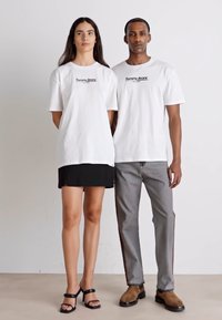 Dos modelos están uno al lado del otro, ambos vistiendo camisetas blancas de gran tamaño con el logo de "Tommy Jeans". La mujer combina la suya con una falda negra y sandalias, mientras que el hombre lleva pantalones grises con franjas laterales y botas marrones.