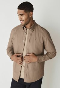 Beige overhemd met puntige kraag, lange mouwen en een logo op de borst, gelaagd over een beige T-shirt, gecombineerd met donkere denim.
