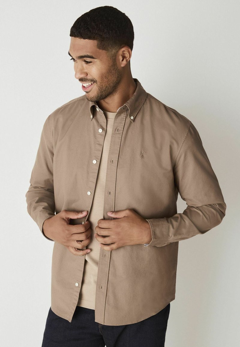 Beige overhemd met puntige kraag, lange mouwen en een logo op de borst, gelaagd over een beige T-shirt, gecombineerd met donkere denim.