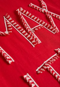 Tela roja con letras bordadas en blanco y acentos texturizados, que incluyen bordes deshilachados y detalles de costura decorativa.