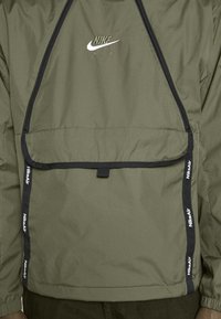 Olivgrön Nike Air-jacka med framficka, svart kant och broderad logotyp. Har justerbara remmar och texturerat material.