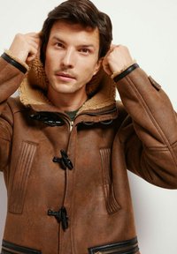 Brązowy skórzany płaszcz z pluszowym, beżowym kołnierzem, z zapięciami na toggle, pionowymi prążkowanymi akcentami i kieszeniami na zamek błyskawiczny.