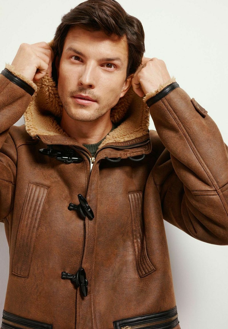 Brauner Ledermantel mit einem plüschigen, beige farbenen Kragen, Knopfverschlüssen, vertikalen Rippenelementen und Reißverschlusstaschen.