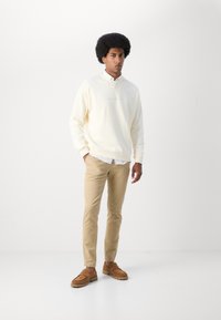 Lichte crème sweatshirt met een ronde hals, beige chino's en bruine instap-schoenen. Model staat tegen een effen achtergrond.