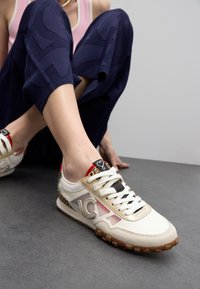 Zapatillas bajas con parte superior en crema y detalles transparentes, estampado de leopardo en el talón, terminadas con una suela de goma y sistema de cordones en la parte delantera.