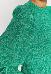 Abito verde con un motivo a pois, caratterizzato da maniche a sbuffo e scollo rotondo, realizzato in un tessuto leggero con una texture liscia.