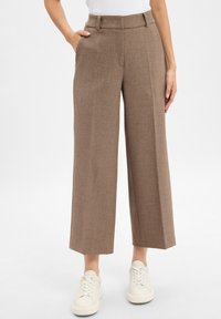 Bruine, wijde broek met een textuurstof, voorzien van een taille met riemlussen en zijzakken, gedragen met witte sneakers.