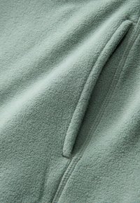 Fleecejakke i lys grøn, med en blød tekstur, en lynlåslomme og sømme langs kanterne, der viser et minimalistisk design.
