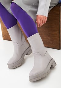Botas altas hasta la rodilla de cuero gris con suela gruesa y parte superior inclinada, combinadas con medias moradas. Textura lisa y diseño minimalista.