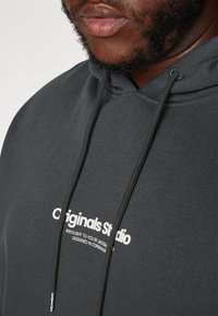 Sudadera gris oscuro hecha de tejido suave. Presenta un bolsillo canguro, capucha con cordón y texto impreso en blanco en el pecho.