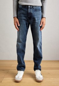 Man wearing dark blue jeans, white sneakers with gum soles, standing on a wooden floor against a plain gray wall.