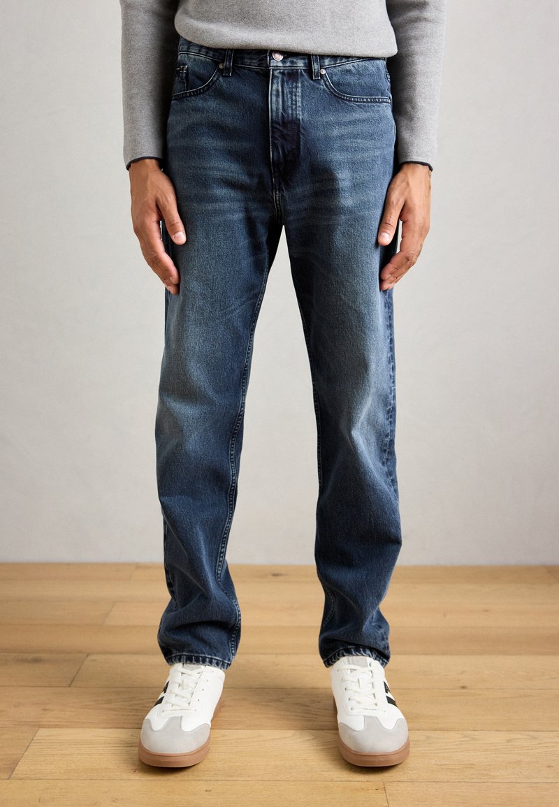 Man wearing dark blue jeans, white sneakers with gum soles, standing on a wooden floor against a plain gray wall.