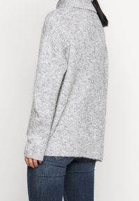 Pull en maille grise avec col montant, coupe oversize et poignets côtelés. Texture douce, présentant de subtiles variations de couleur et de motif.