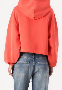 Sudadera corta coral con capucha, mangas oversize y puños acanalados, combinada con vaqueros azul claro que muestran los bolsillos traseros.