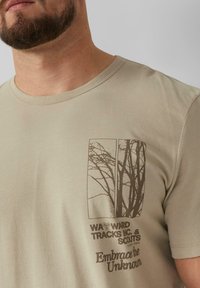 Beige katoenen t-shirt met een boom silhouet grafiek in donkerbruin aan de linkerkant, met de tekst "Omarm het Onbekende." Rond de hals, korte mouwen.