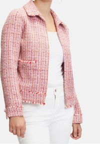 Różowa i biała tweedowa kurtka z kołnierzykiem, teksturowanym wykończeniem i frędzlami na brzegach. Posiada dwie przednie kieszenie oraz zapięcie na guziki.