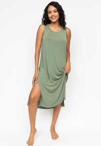 Robe sans manches verte en tissu doux, avec une coupe décontractée, une fente sur le côté et un design drapé. Pieds nus visibles sur l'image.