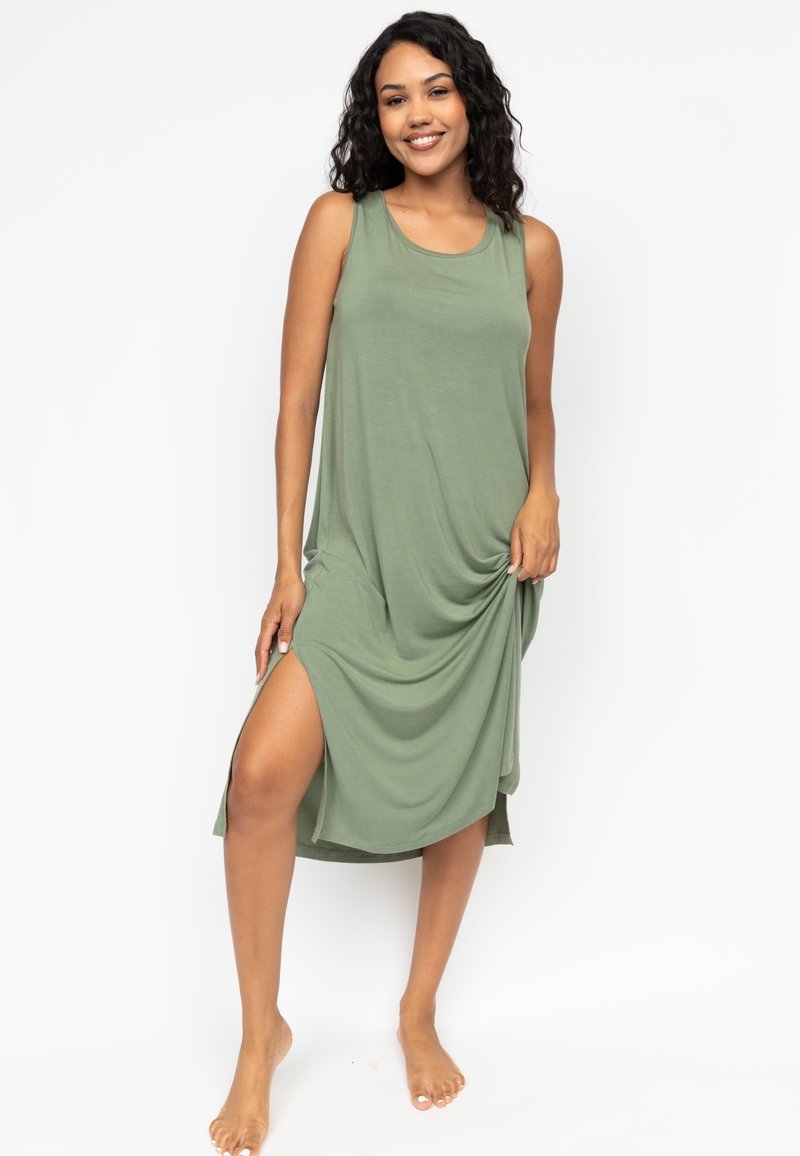 Robe sans manches verte en tissu doux, avec une coupe décontractée, une fente sur le côté et un design drapé. Pieds nus visibles sur l'image.