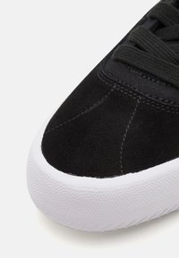 Adidași negri cu un superior din piele întoarsă, având o suprafață texturată, talpă din cauciuc alb și sistem de șireturi negre.