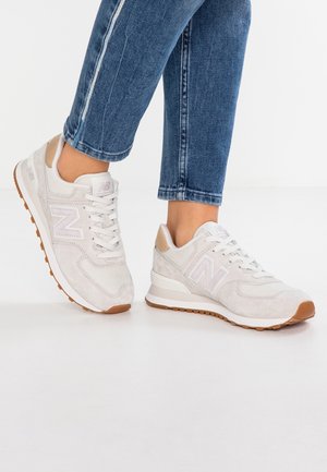Personne portant des baskets New Balance beige clair avec des lacets blancs et des semelles en gomme, assorties à un jean en denim bleu coupé au-dessus de la cheville.