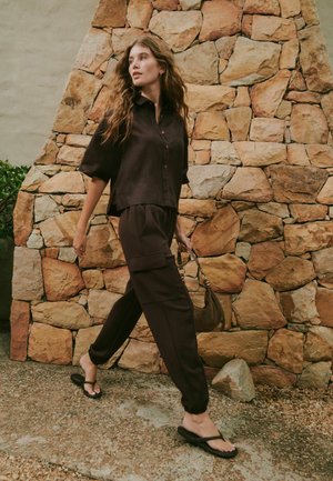 Frau mit langen, welligen Haaren, die an einer Steinmauer vorbeigeht und ein dunkles, locker sitzendes Hemd, Cargohosen, Flip-Flops trägt und eine kleine braune Handtasche hält.