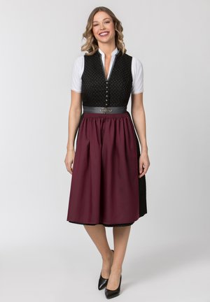 Lächelnde Frau, die ein traditionelles schwarzes Dirndl mit weißer Bluse und dunkelroter Schürze trägt, steht mit überkreuzten Füßen vor grauem Hintergrund.